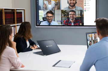 Image of a group of three people sitting at a conference table with a Dynabook laptop computer; the people are focusing on a video conference call displayed on a Windows Collaboration Display from Sharp unit mounted on the wall ahead of them